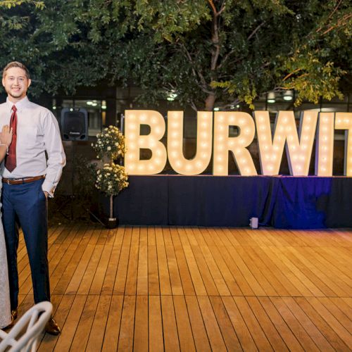 A prom couple stands by a glowing &ldquo;BURWITZ&rdquo; sign at an outdoor event, under string lights and greenery.
