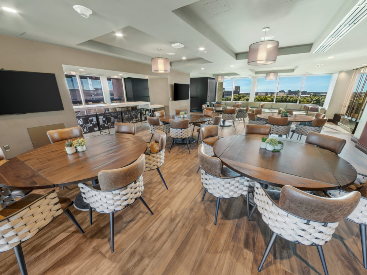 A modern conference room with wooden tables, woven chairs, pendant lights, TV screens, and large windows overlooking the outdoors.