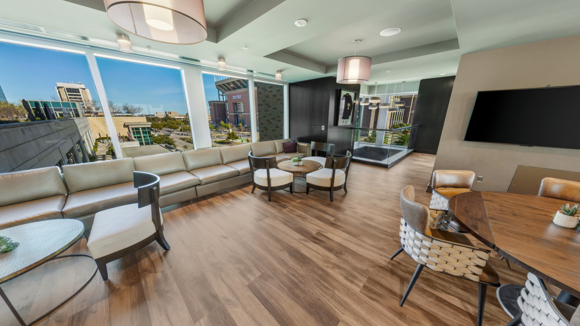Bright modern lounge with leather sofas and round table, floor-to-ceiling windows, outdoor view, and an HD TV on the wall.