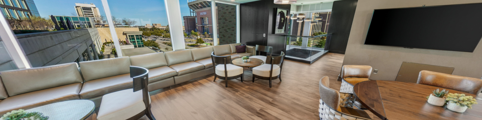 Bright modern lounge with leather sofas and round table, floor-to-ceiling windows, outdoor view, and an HD TV on the wall.