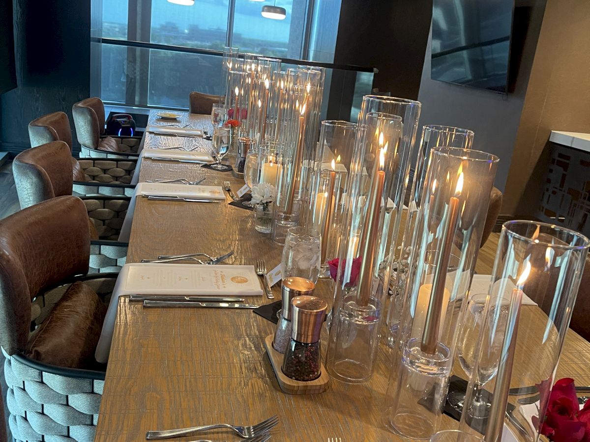 A neatly set dining table for a celebration with glowing candles, glassware, menus, and cushioned chairs in a modern room.