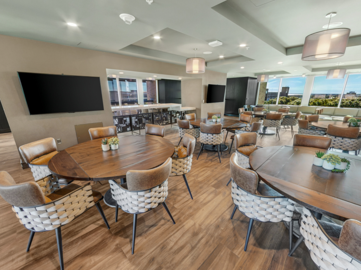 Modern conference lounge with round wooden tables, upholstered chairs, a TV wall, and large windows overlooking a city.