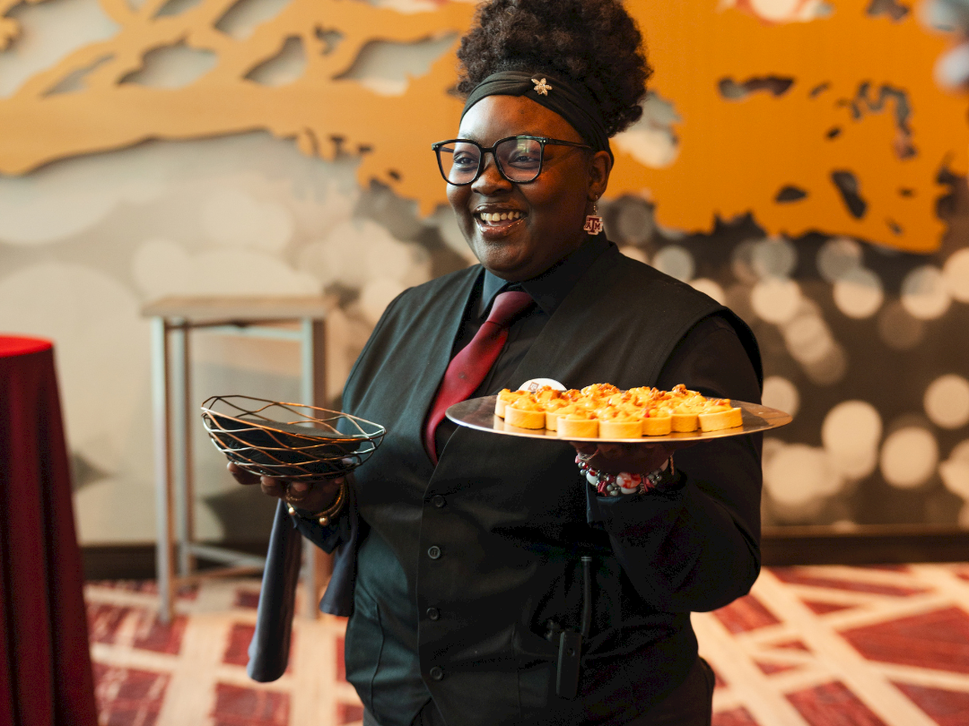 A smiling woman in a suit holds a tray of bite-sized food at an event with a festive orange backdrop.