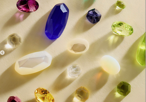 A spread of colorful faceted gemstones&mdash;purple, blue, green, yellow, and red&mdash;scattered on a light background.