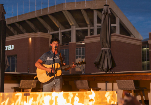 An outdoor musician sings and plays acoustic guitar beside a roaring fire under a large pavilion, night-time.