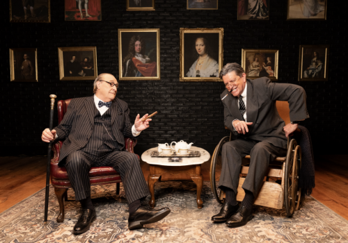 Two men in a cozy room play-act tea-time: one in a wheelchair pours, the other sits chatting beside a teapot and cups.