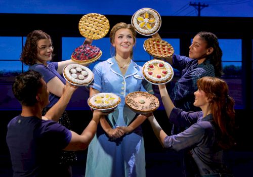 A contestant smiles center stage as helpers surround her with colorful pies and waffles on trays in a blue studio, everyone grins.