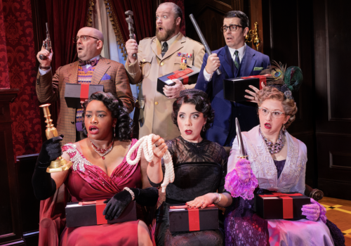 A comedic troupe in vintage costumes on a theater stage holds prop guns and gifts in red-and-gold boxes.