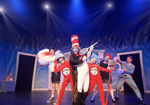 A circus troupe in red-and-white outfits dances under blue spotlights, with a clown holding a tennis racket and a bear-costumed performer.
