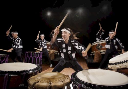A group of drummers in matching black-and-white outfits performs with sticks and large taiko drums under bright stage lights.