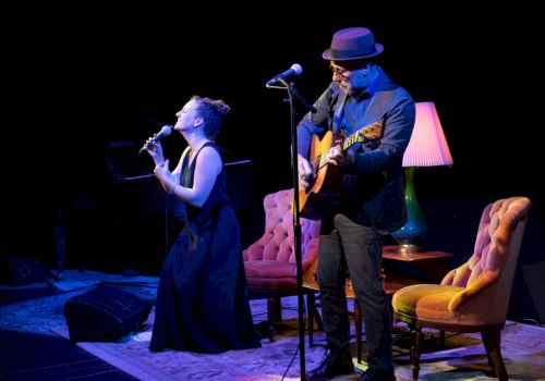 A live music performance on stage: a singer at a microphone and a guitarist in a hat playing beside him, under purple lights.