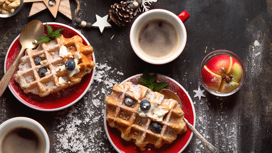 Two plates of waffles with blueberries, accompanied by cups of coffee, and festive decorations on a table with cinnamon and a bowl of nuts.