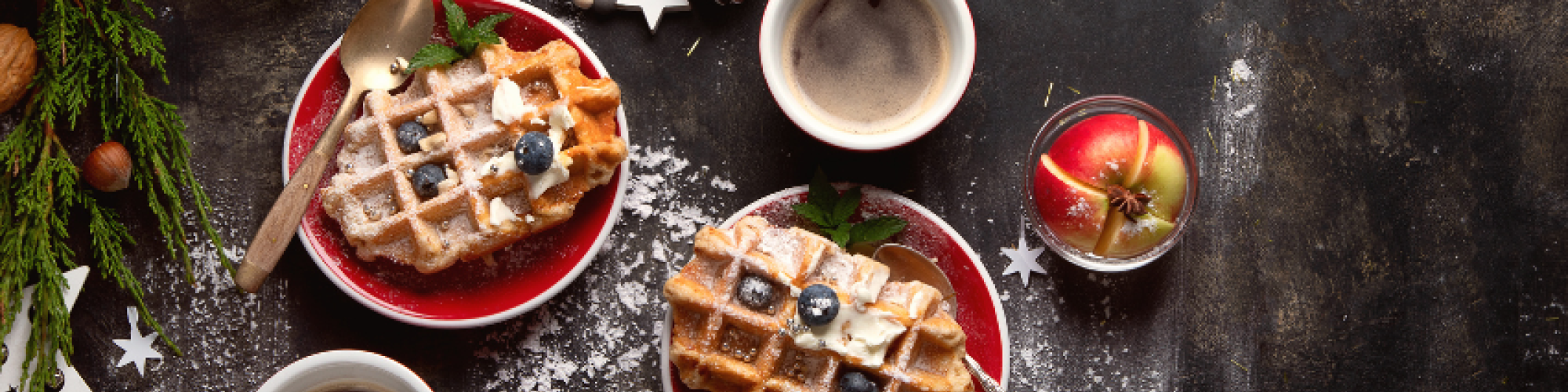 Two plates of waffles with blueberries, accompanied by cups of coffee, and festive decorations on a table with cinnamon and a bowl of nuts.