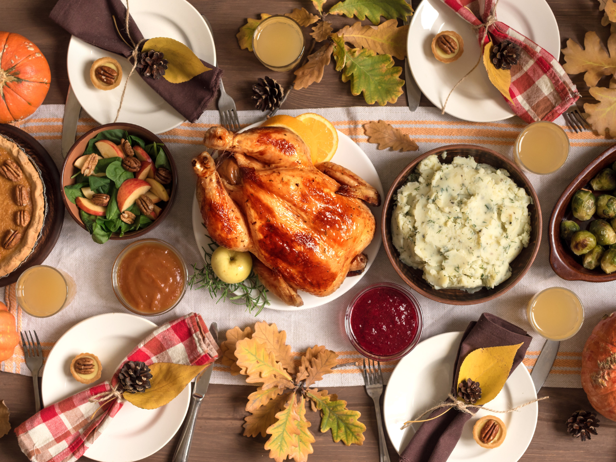 A Thanksgiving meal setup with roast turkey, mashed potatoes, cranberry sauce, Brussels sprouts, salad, pie, and drinks, surrounded by fall decor.