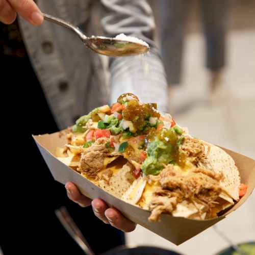 Street tacos topped with chopped cilantro, onions, and salsa verde in a takeaway tray, being scooped with a spoon.