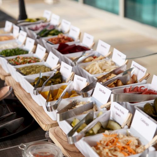 A colorful salad bar with assorted toppings in labeled containers, ready for guests to customize their bowls, on a sunny outdoor setup.