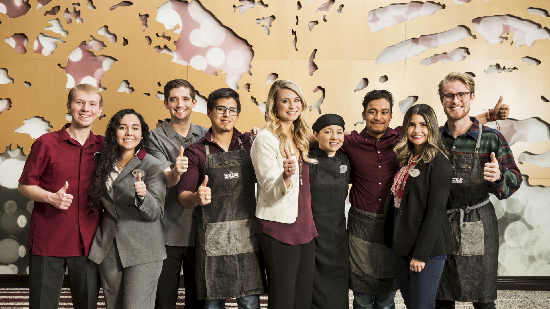 A group of people giving thumbs up for a photo. Some wear aprons and some wear business attire, standing in front of an artistic wall.