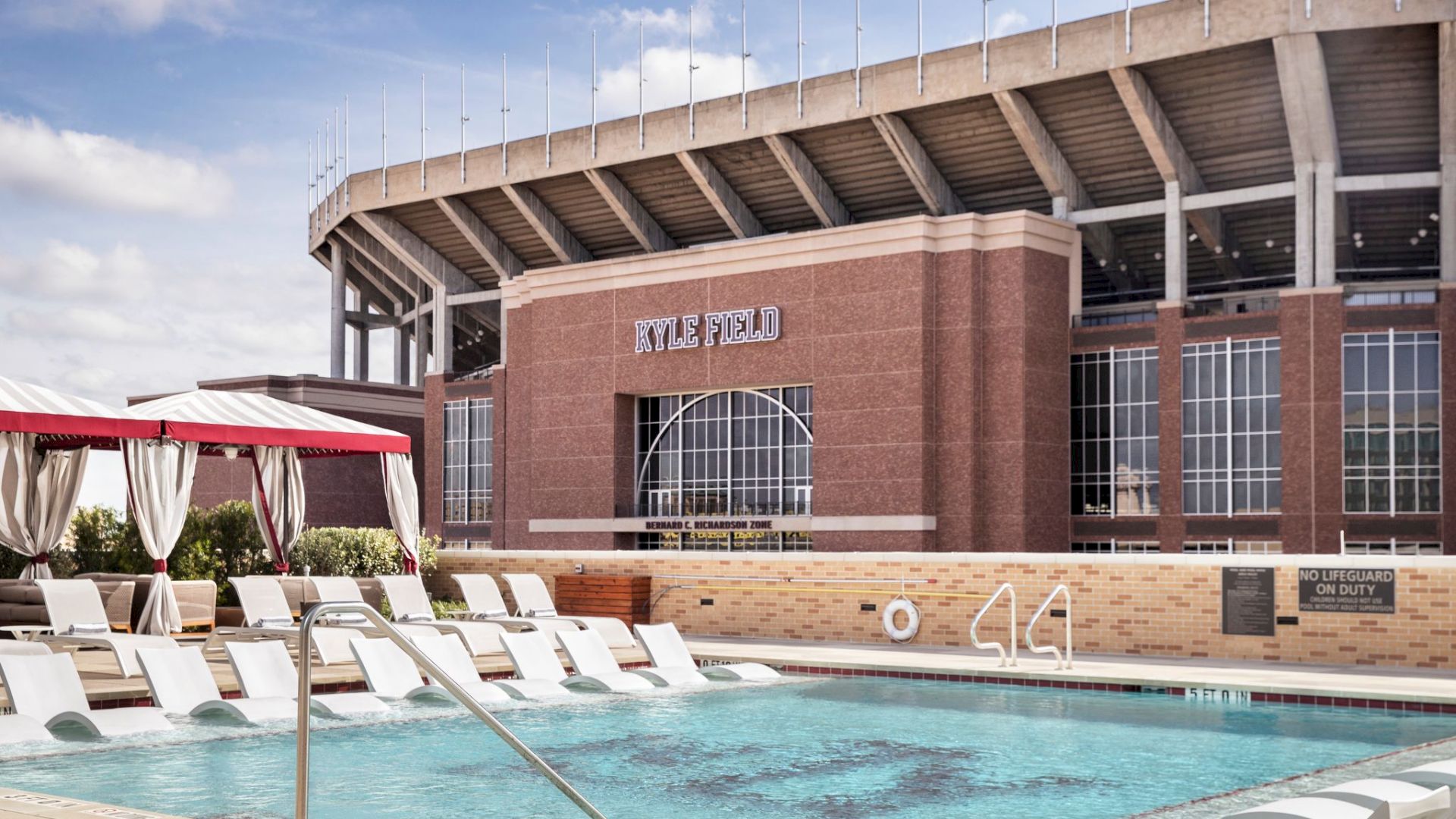 This image shows a swimming pool with lounge chairs, a cabana, and a large stadium named Kyle Field in the background.