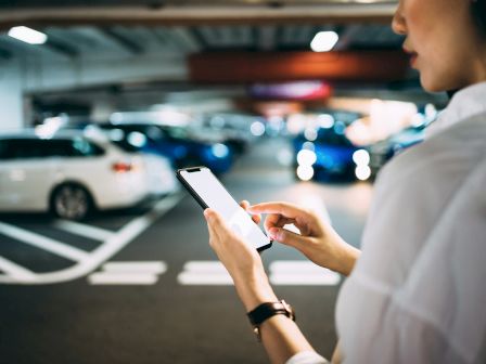 A person holds a smartphone in a bright indoor venue while people move around in the background, looking like an airport or station.