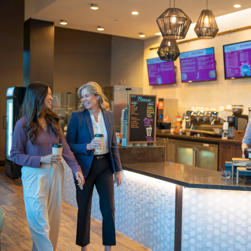 Two women chat with coffee cups in a modern caf&eacute; by the barista counter and digital menu screens.