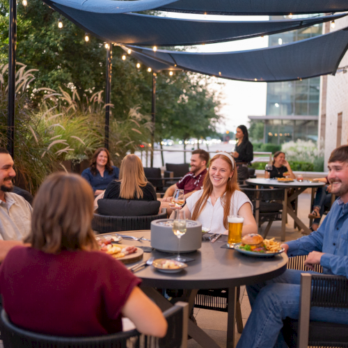 Friends dine outdoors at a patio table under blue awnings and warm string lights, laughing over drinks and food.