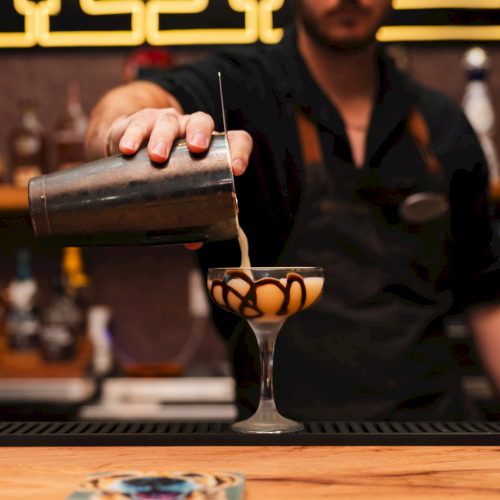 A bartender pours a creamy coffee-like drink from a metal shaker into a martini glass in a cozy bar.