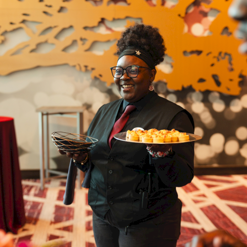 A smiling woman in a vest holds a platter of orange-topped appetizers at an event with a patterned wall behind her.
