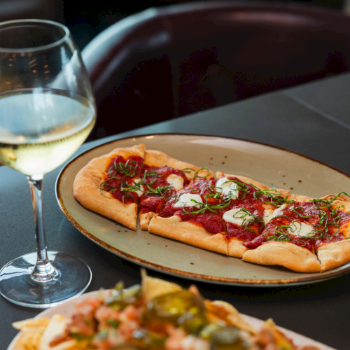 A thin-crust pizza topped with mozzarella, pepperoni, sauce, and herbs, beside a glass of white wine.