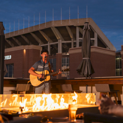 A young musician sings and plays acoustic guitar beside a roaring campfire at an outdoor event.