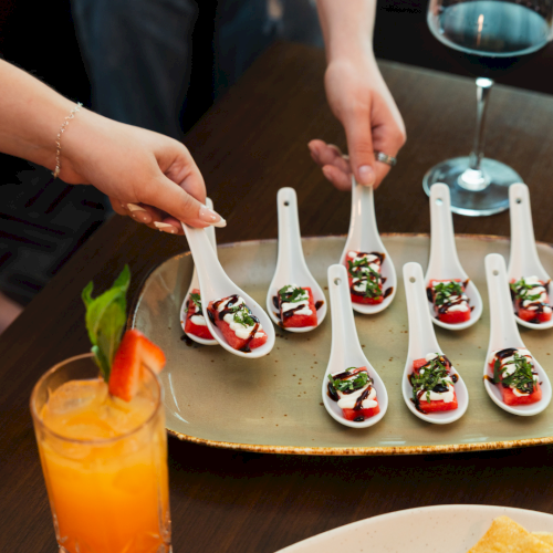 A chef&rsquo;s hands arrange bite-size appetizers on white spoons, topped with sauce and herbs, on a tray at a table.