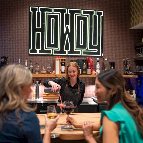 A bartender pours drinks for guests in a cozy bar with a glowing neon sign on the wall.