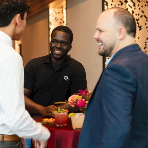 A smiling man in a black polo chats at an event table with colorful flowers and drinks.