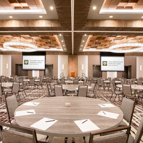 A modern conference room with round tables, chairs, and large screens displaying Texas A&M Hotel and Conference Center.