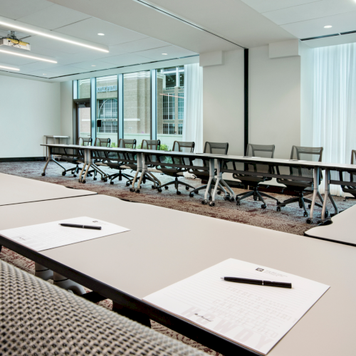 A modern conference room with long tables, rolling office chairs, ceiling lights, and windows showing a city building through curtains.