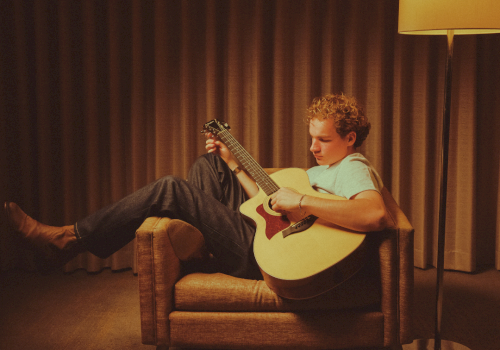 A boy sits on a couch playing an acoustic guitar, with warm lamp light and curtains behind him.