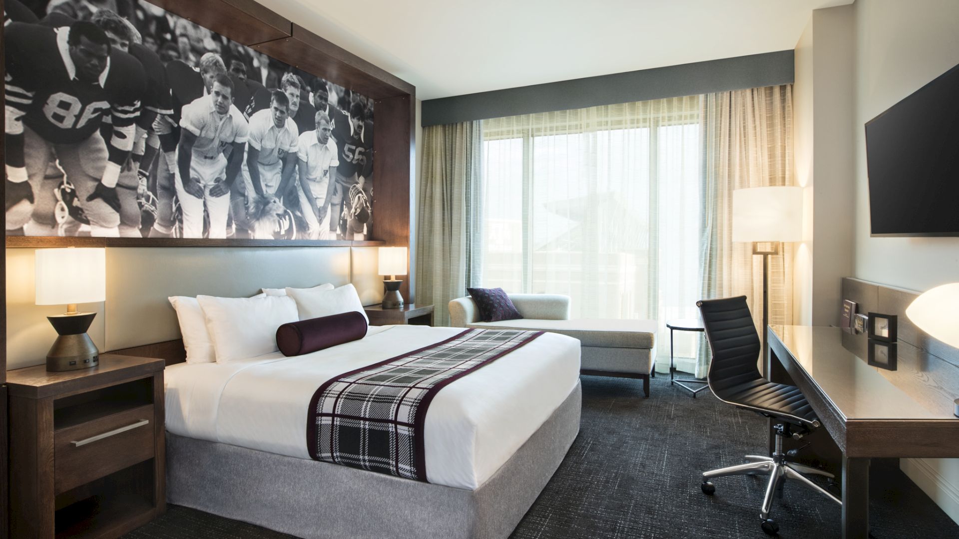 The image shows a modern hotel room with a large bed, a sports-themed picture, a desk, a TV, and a window with sheer curtains.