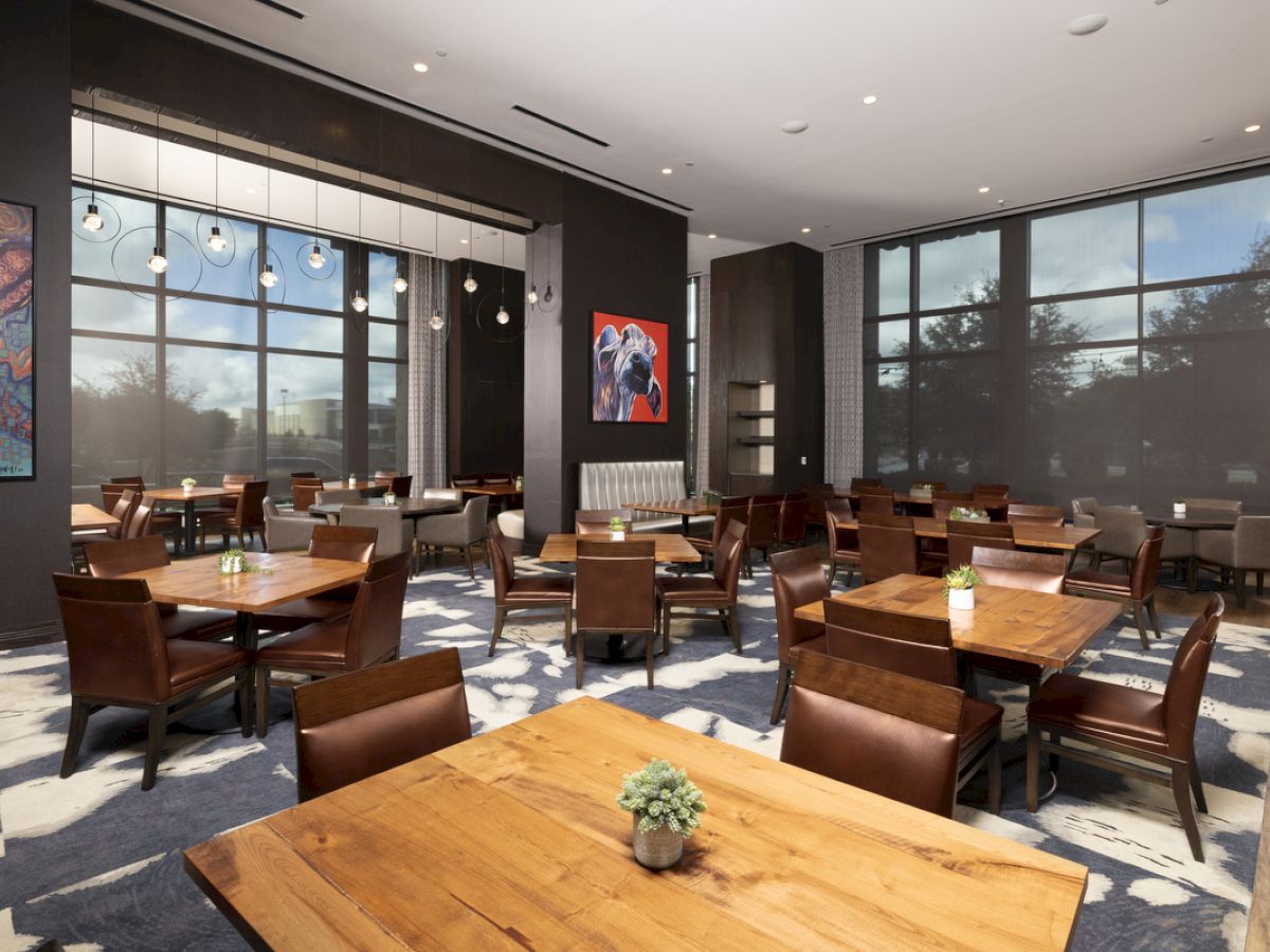 The image shows a modern dining area with wooden tables and chairs, large windows, abstract paintings, and a lush carpet.