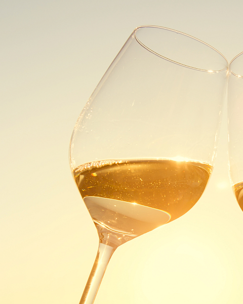 Two stemmed wine glasses filled with amber-gold liquid, catching warm golden light against a pale background.