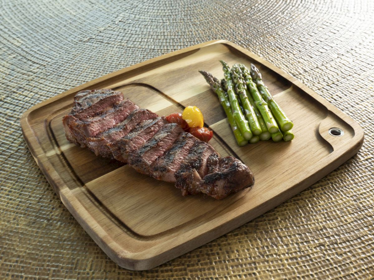 A grilled steak with asparagus on a wooden tray, plus a small tomato garnish, photographed on a textured surface.