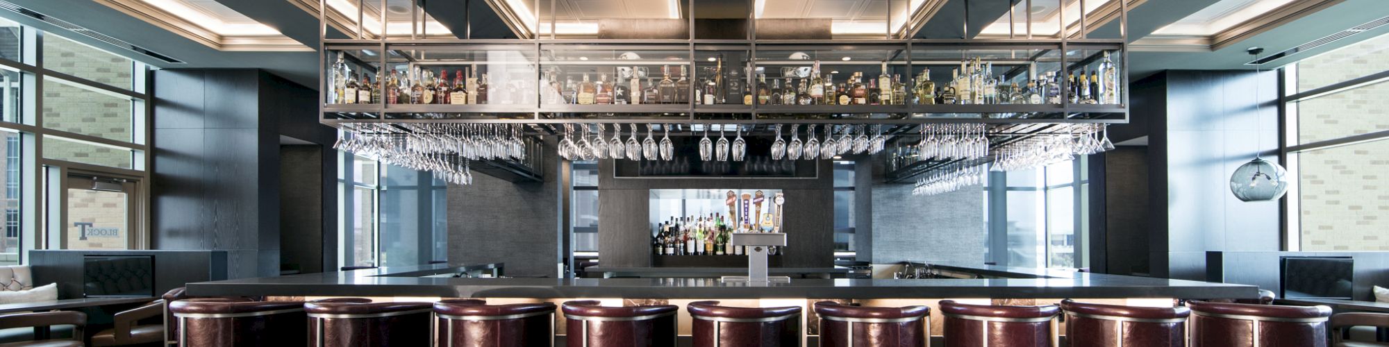 A modern bar with high stools, hanging glasses, bottles on display, and a sleek ceiling design, creating a stylish atmosphere.