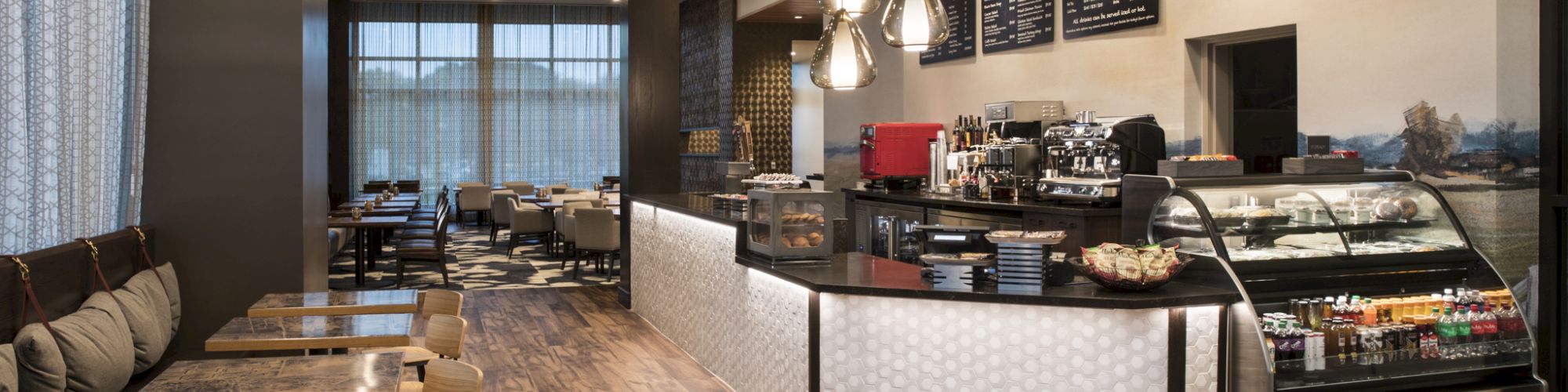 The image shows a modern caf&eacute; interior with a wooden floor, tables, cushioned seating, a counter with drinks, and pastries on display.