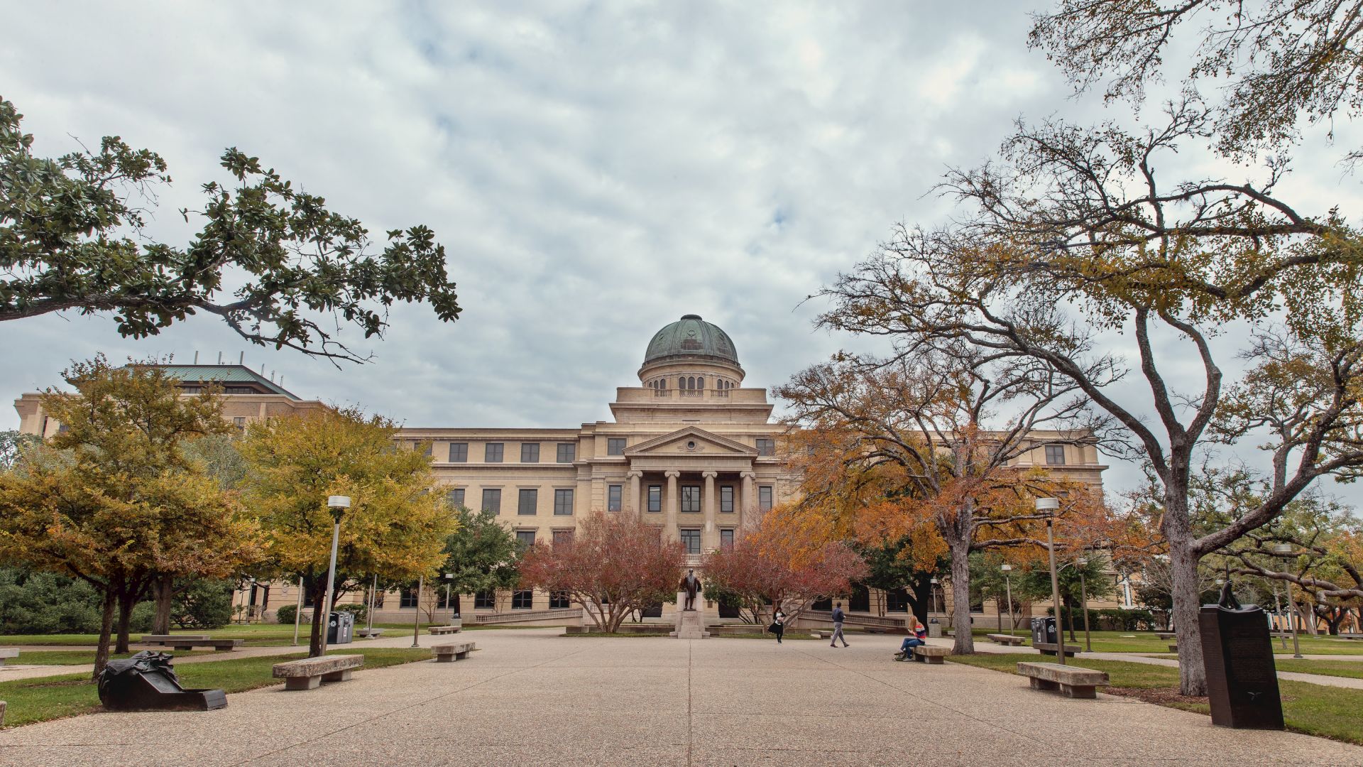 A university courtyard with a grand building and central dome, framed by autumn trees under a cloudy sky.