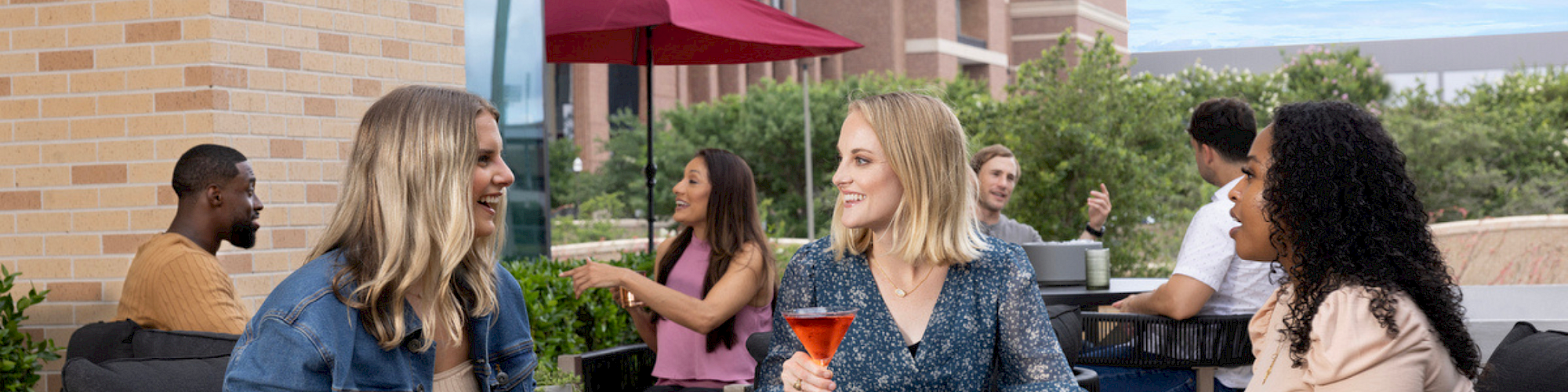 Drafting a sunny rooftop hangout: friends chat and sip colorful cocktails around a round table, with plants and red umbrellas in the background.