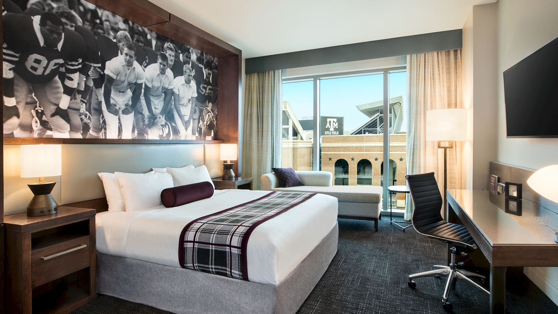 A hotel room with a large bed, art of football players, a desk, and a view of a stadium through a window is shown.