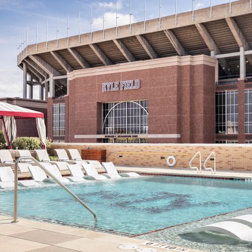 A pool with lounge chairs, a red and white cabana, and a large brick stadium in the background. The stadium says "Kyle Field."