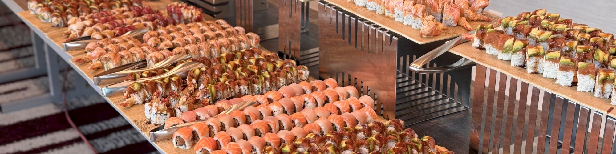 A sushi buffet with rows of maki and nigiri&mdash;salmon, tuna, and shrimp&mdash;served on wooden trays by a catering station.