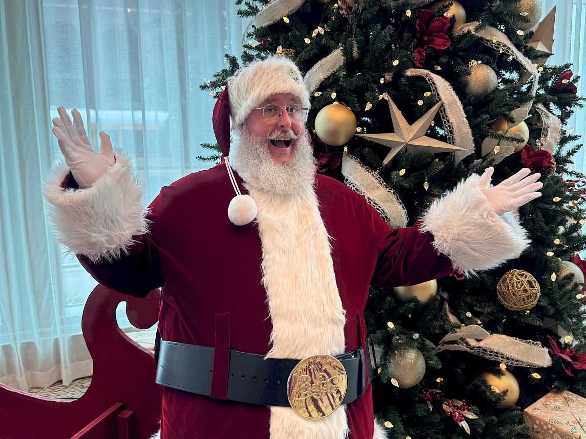 Santa in a red suit with a white beard waves beside a decorated Christmas tree with ornaments and gifts.